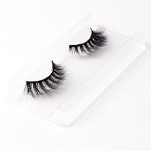 Könnte beinhalten: Ein Paar schwarze, voluminöse falsche Wimpern auf einer transparenten Kunststoffschale. Die Wimpern sind dicht mit einzelnen Strähnen gefüllt, wodurch ein voller und dramatischer Effekt entsteht. Die Schale hat ein schlichtes, klares Design, das die Wimpern hervorhebt.