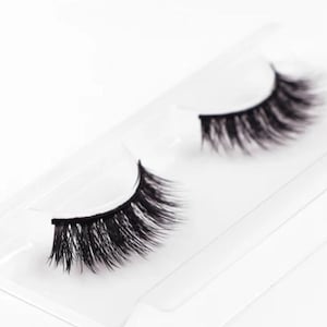 Könnte beinhalten: Ein Paar schwarze falsche Wimpern, präsentiert in einer transparenten Kunststoffverpackung. Die Wimpern sind dicht mit einzelnen Strähnen gepackt, wodurch ein voller und voluminöser Look entsteht. Die Verpackung ist schlicht und sauber.