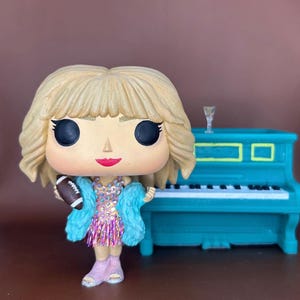 May include: A Funko Pop vinyl figure of a blonde woman with a football, wearing a pink dress, blue fur coat, and pink boots. A teal piano is to the right. The figure has large black eyes and a small red mouth.