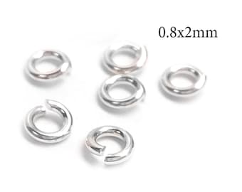 Puede incluir: Cinco pequeñas anillas de salto de metal plateado con forma circular y una abertura visible en un lado. Las anillas están dispuestas de forma dispersa y el texto 0.8x2mm está impreso en negro en la esquina superior derecha.