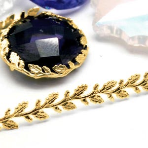May include: A gold-colored leaf-shaped trim and a dark blue faceted gemstone with a gold leaf-shaped bezel. The trim is a repeating pattern of leaves. The gemstone is round and has a faceted surface.