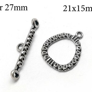 Könnte beinhalten: Zwei silberfarbene Metallverschlüsse zum Schmuckbasteln. Einer ist eine 27 mm lange Stange mit einem Perlmuster. Der andere ist ein 21 x 15 mm großes Oval mit einem Perlmuster.