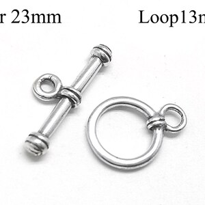 Könnte beinhalten: Ein silberfarbenes Knebelverschluss-Set. Der Stab misst 23 mm, und die Schlaufe 13 mm. Der Verschluss ist für die Schmuckherstellung konzipiert, mit einem T-Stab und einer kreisförmigen Schlaufe für sicheren Halt. Das Metall hat eine polierte Oberfläche.