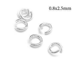 Puede incluir: Una colección de pequeñas anillas de salto de metal plateado con una abertura. Las anillas están dispuestas de forma dispersa y el texto 0.8x2.5mm es claramente visible en la esquina superior derecha, indicando las dimensiones.