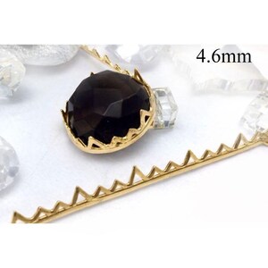 Pode incluir: Um componente de joalharia em metal dourado com uma pedra preciosa castanha escura, com cerca de 4,6 mm. A pedra preciosa está engastada numa cravação decorativa em forma de coroa. Também são visíveis peças metálicas douradas adicionais com um design em ziguezague.