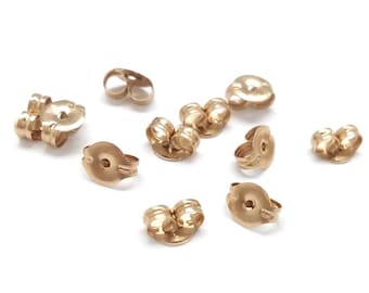 May include: A collection of gold-toned metal earring backs, each featuring a circular disc with a central hole and two curved, spring-loaded tension loops designed to securely grip an earring post. The set includes multiple individual pieces.