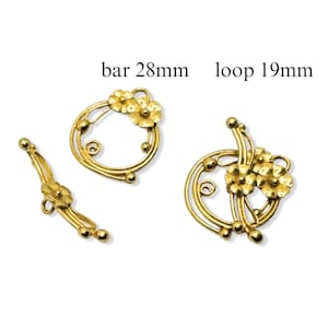 Könnte beinhalten: Zwei goldfarbene Metall-Schmuck-Findings mit Blumenmotiven. Der größere Finding ist eine 19 mm Schleife mit zwei Blumen. Der kleinere Finding ist eine 28 mm Stange mit zwei Blumen.