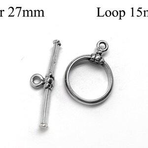 Könnte beinhalten: Silberner Toggle-Verschluss mit einer 27 mm langen Stange und einer 15 mm großen Schlaufe. Die Stange hat an einem Ende eine kleine Schlaufe und am anderen Ende einen dekorativen Knoten. Die Schlaufe ist rund und hat oben einen dekorativen Knoten.