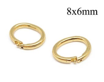 Könnte beinhalten: Zwei goldfarbene Metall-Biegeringe mit einem aufklappbaren Verschlussmechanismus. Jeder Ring verfügt über einen kleinen hervorstehenden Stift und ein entsprechendes Loch zum Schließen. Der Text 8x6mm ist schwarz über den Ringen abgedruckt.