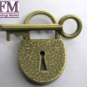 Puede incluir: Un candado de latón de estilo vintage con una llave adjunta. El candado tiene un acabado martillado y una cerradura en el centro. La llave está unida al candado con un anillo.