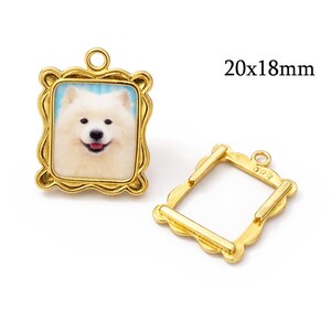 Könnte beinhalten: Goldfarbener Metallanhänger mit einem Bild eines lächelnden weißen Hundes in einem dekorativen Rahmen. Der Rahmen misst 20x18mm. Der Anhänger hat oben eine Schlaufe zum Befestigen an Schmuck oder anderen Bastelarbeiten.