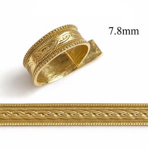 以下が含まれることがあります： 複雑な編み込み模様のゴールドカラーのリング、幅7.8mm。リングは白い背景に表示されています。画像には、お揃いの編み込みトリムの一部も表示されています。リングの幅は約0.8cmです。