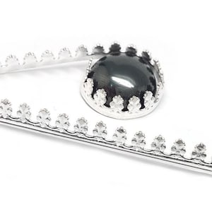 May include: A silver-toned metal brooch featuring a long, straight pin with a decorative scalloped edge. A circular, polished black cabochon stone is securely set within a matching silver-toned crown-style bezel setting attached to the pin.