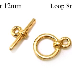 Könnte beinhalten: Goldfarbener Metallverschluss mit einem 12 mm langen Steg und einer 8 mm großen Schlaufe. Der Steg hat an einem Ende eine kleine Schlaufe und in der Mitte ein gedrehtes Design. Die Schlaufe hat oben ein gedrehtes Design.