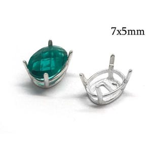 Pode incluir: Uma pedra preciosa oval facetada verde-esmeralda num engaste de metal prateado com garras, ao lado de um engaste de metal prateado vazio a condizer. O texto 7x5mm é visível, indicando as dimensões dos componentes de engaste ovais.