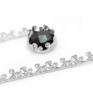 May include: A silver metal crown-shaped setting with a dark green gemstone. The setting features an ornate, swirling design. A separate piece of silver metal with a similar design is also visible.