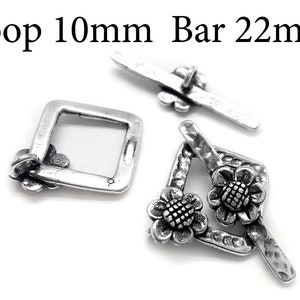 Könnte beinhalten: Silberfarbener Metallverschluss mit einer 10 mm Schlaufe und einer 22 mm Stange. Der Verschluss hat ein dekoratives Blumenmuster.