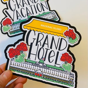 May include: A black and white illustration of the Grand Hotel on Mackinac Island. The illustration features a yellow awning with the text "Mackinac Island" and the words "Grand Hotel" in black lettering. The illustration is surrounded by red and green flowers.