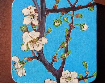 May include: Square coaster with a painting of white blossoms and green buds on a brown branch against a bright blue background. The coaster has rounded corners and is likely made of wood.