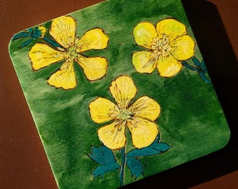 May include: Square wooden coaster with a green background and three yellow buttercup flower illustrations. The flowers have detailed yellow petals and green and blue stems and leaves. The coaster is a decorative item.