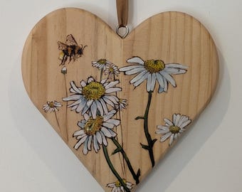 May include: Wooden heart-shaped wall hanging with a painted floral design. The artwork features white daisies with yellow centers and a bee. A brown ribbon is attached for hanging. The background is a light wood grain.