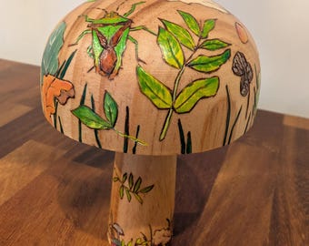May include: A wooden mushroom sculpture with a painted design. The mushroom cap features a green and brown insect, green leaves, and other plant life. The stem is also painted with leaves and small mushrooms. The sculpture is on a wooden surface.