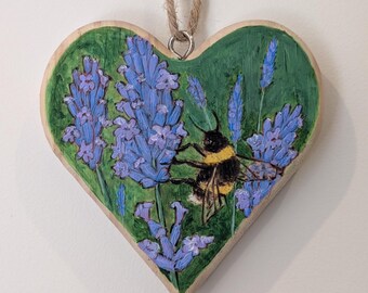 May include: Wooden heart-shaped ornament featuring a painted scene of lavender and a bumblebee. The background is green, and the lavender is a vibrant purple. A natural twine loop is attached for hanging.