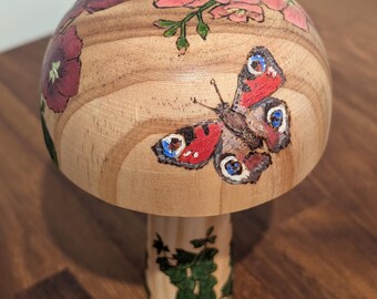 May include: A wooden mushroom-shaped decorative item. The top is painted with pink and purple flowers and a butterfly with red, blue, and black markings. The stem features green painted foliage. The item is made of light-colored wood.