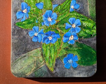 May include: Square wooden coaster featuring a painting of blue flowers with white centers and green leaves. The background is a dark gray with a textured appearance. The coaster is a decorative item for home use.