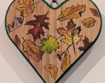 May include: Wooden heart-shaped ornament with a green border and a brown ribbon for hanging. The ornament is decorated with painted autumn leaves in shades of yellow, orange, red, and green, creating a seasonal theme.