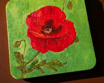 May include: Square wooden coaster featuring a vibrant red poppy flower against a green background. The poppy has detailed petals and a dark center. Green leaves and buds accompany the flower, creating a natural theme. The coaster is set on a brown surface.