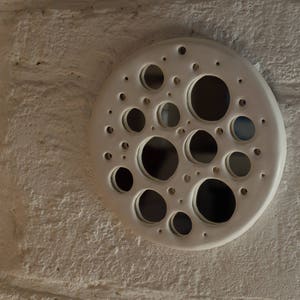 Op de afbeelding: Witte keramische wanddecoratie met een cirkelvormig ontwerp met meerdere uitgesneden cirkels in verschillende maten.