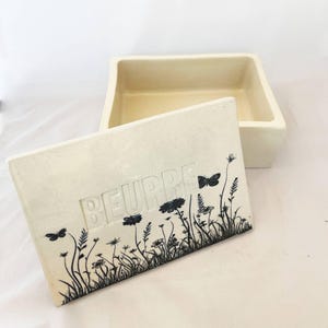 Könnte beinhalten: Eine cremefarbene Butterdose aus Keramik mit rechteckigem Unterteil und passendem Deckel. Auf dem Deckel befindet sich das geprägte Wort BEURRE sowie eine schwarze botanische Illustration von Wildblumen und Schmetterlingen am unteren Rand.
