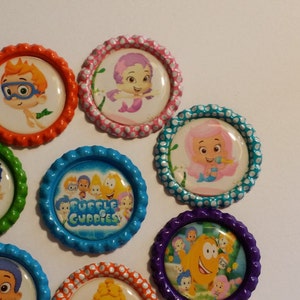 May include: Eight colorful round bottle cap stickers featuring characters from the animated television series Bubble Guppies. The stickers feature the characters Goby, Molly, Gil, Deema, Oona, Nonny, and the Bubble Guppies logo.