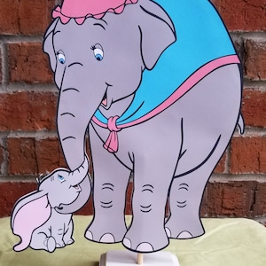 May include: A gray mother elephant with a pink bonnet and blue cape holding her gray baby elephant. The mother elephant has a pink bow on her bonnet. The baby elephant has pink ears. The elephants are standing on a white square base.