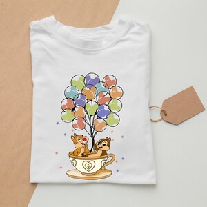 May include: White t-shirt with a cartoon image of Chip and Dale from Disney's Chip 'n Dale Rescue Rangers. The chipmunks are holding onto a bunch of colorful balloons in the shape of Mickey Mouse heads. They are sitting in a teacup with a heart design.