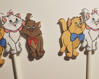 May include: Three cartoon cat cupcake toppers. The cats are from the Disney movie The Aristocats. The cats are a white cat with a pink bow, a brown cat with a red bow, and a yellow cat with a blue bow.