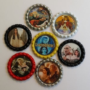 May include: A collection of seven bottle cap magnets featuring images from the movie "Labyrinth." The caps have various colored rims, including black, red, silver, white, and yellow. The images depict scenes and characters from the film.