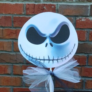 Pu&ograve; includere: Un ritaglio di carta bianca del viso di Jack Skellington con occhi neri e una bocca cucita. Il ritaglio &egrave; attaccato a un bastone con un fiocco di tulle bianco.