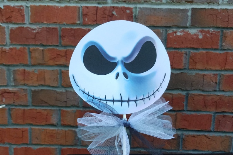 Pu&ograve; includere: Un ritaglio di carta bianca del viso di Jack Skellington con occhi neri e una bocca cucita. Il ritaglio &egrave; attaccato a un bastone con un fiocco di tulle bianco.