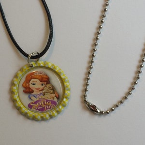 May include: A round pendant necklace featuring the Disney character Sofia the First, with a yellow scalloped edge. The pendant is attached to a black cord. A separate silver ball chain necklace is also present.