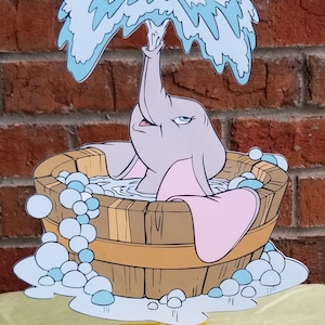 May include: A cutout of a gray elephant taking a bath in a wooden tub with blue and white bubbles. The elephant is holding a blue and white water spout above its head. The cutout is on a yellow stand.