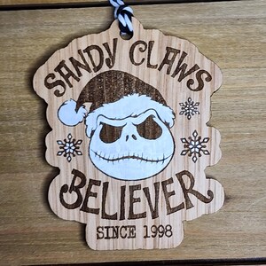 May include: A wooden Christmas ornament featuring a cartoon character with a Santa hat and the text "Sandy Claws Believer Since 1998".