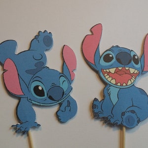 Puede incluir: Dos adornos para tartas de Stitch azul con orejas rosas. Un Stitch est&aacute; de pie sobre su cabeza con los brazos levantados y el otro Stitch est&aacute; sentado con las piernas cruzadas.