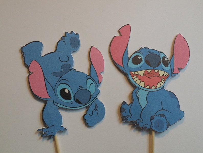 Puede incluir: Dos adornos para tartas de Stitch azul con orejas rosas. Un Stitch est&aacute; de pie sobre su cabeza con los brazos levantados y el otro Stitch est&aacute; sentado con las piernas cruzadas.