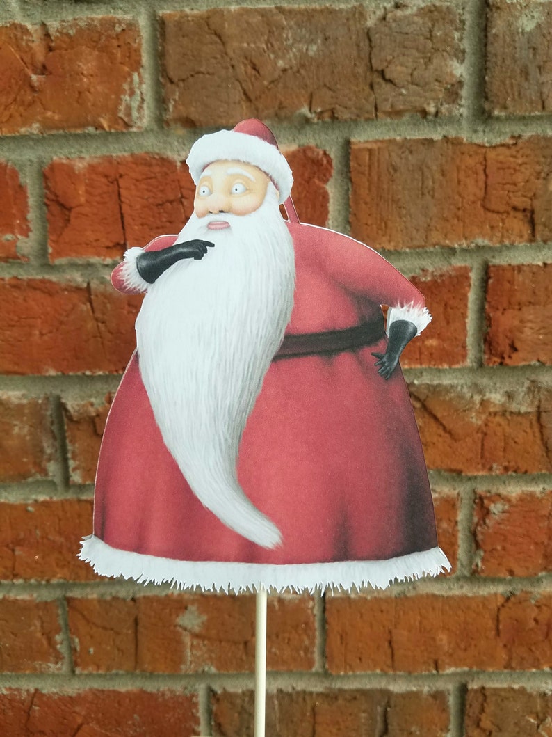 May include: A red and white Santa Claus cutout with a white beard and black gloves. The cutout is on a stick and is in front of a brick wall.