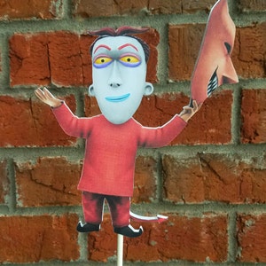 May include: A cutout of a character from The Nightmare Before Christmas, dressed in red with a devil tail, holding a mask. The character has pale skin, purple eye shadow, and yellow eyes. The background is a red brick wall.