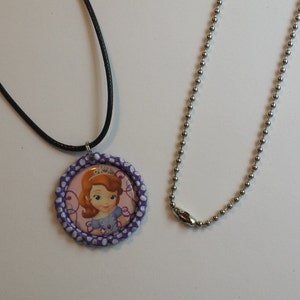 May include: A round pendant necklace featuring a cartoon image of a princess with a tiara, set in a purple and white polka dot border. The pendant hangs from a black cord necklace. A separate silver ball chain necklace is also shown.