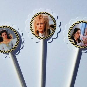 May include: Three round cupcake toppers with black and yellow chevron patterns. The toppers feature images of characters from the movie Labyrinth: a woman with long brown hair, a man with long blonde hair, and a woman holding a baby.