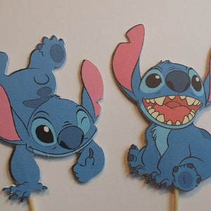 Puede incluir: Dos adornos para tartas de Stitch azul con orejas rosas. Un Stitch est&aacute; de pie sobre su cabeza con el brazo levantado, el otro Stitch est&aacute; sentado con la boca abierta.